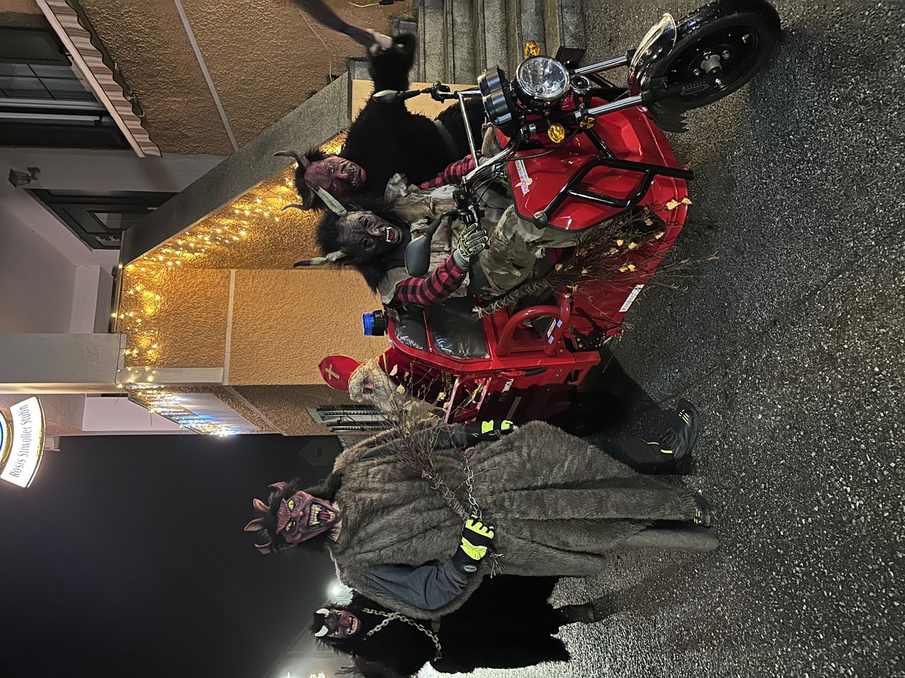 Zwei Menschen in Teufelskostümen posieren neben einem roten Motorrad vor einem Gebäude mit gelben Lichtern in der Nacht.