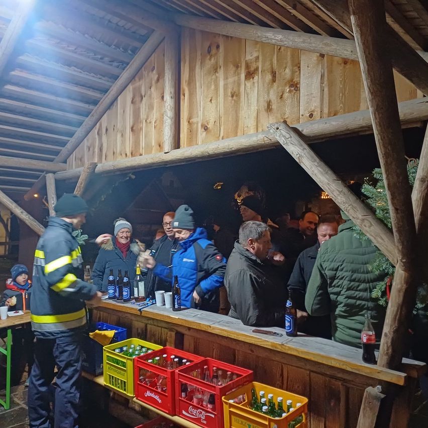 Eine Gruppe von Menschen versammelt sich an einer Bar in einer Holzstruktur. Sie tragen Winterkleidung. Flaschen, Tassen und ein Weihnachtsbaum stehen auf der Theke.