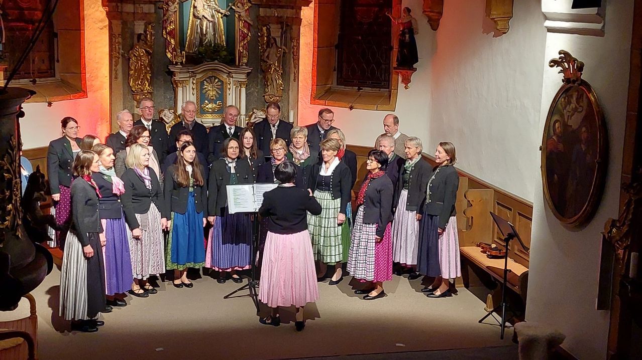 Ein Chor in traditioneller Kleidung steht auf einer Bühne, begleitet von einem Geiger, in einer Kirche mit kunstvollen Dekorationen und religiösen Symbolen.