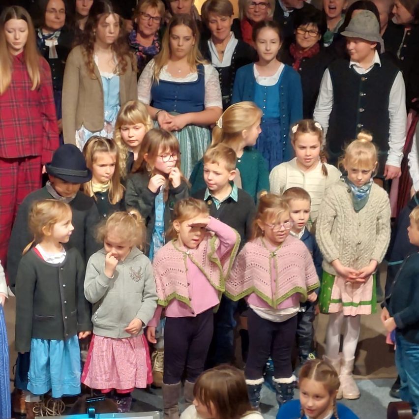 Eine Gruppe von Kindern in Winterkleidung steht zusammen. Einige tragen Brillen, Mützen und Stiefel. Sie scheinen auf etwas zu warten und nach links zu schauen.