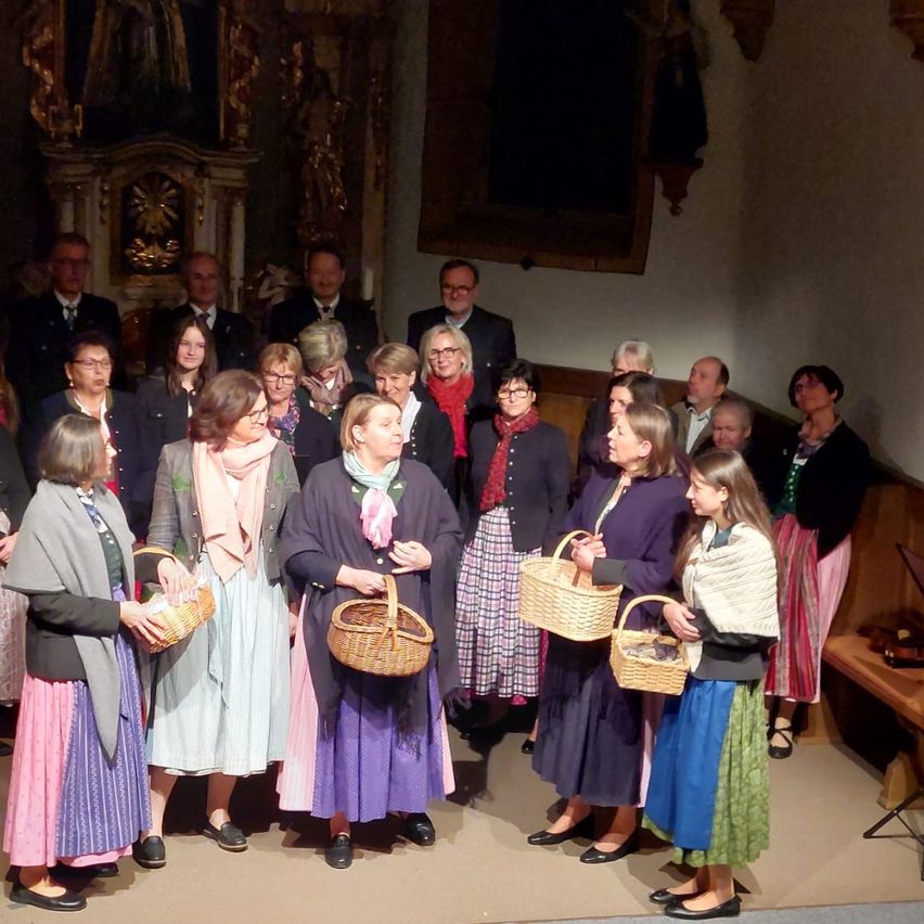 Eine Gruppe von Menschen in traditioneller Kleidung steht in einer Kirche und hält Körbe. Dahinter sitzen andere in formeller Kleidung. Es gibt einen dekorativen Altar und ein Gemälde an der Wand.