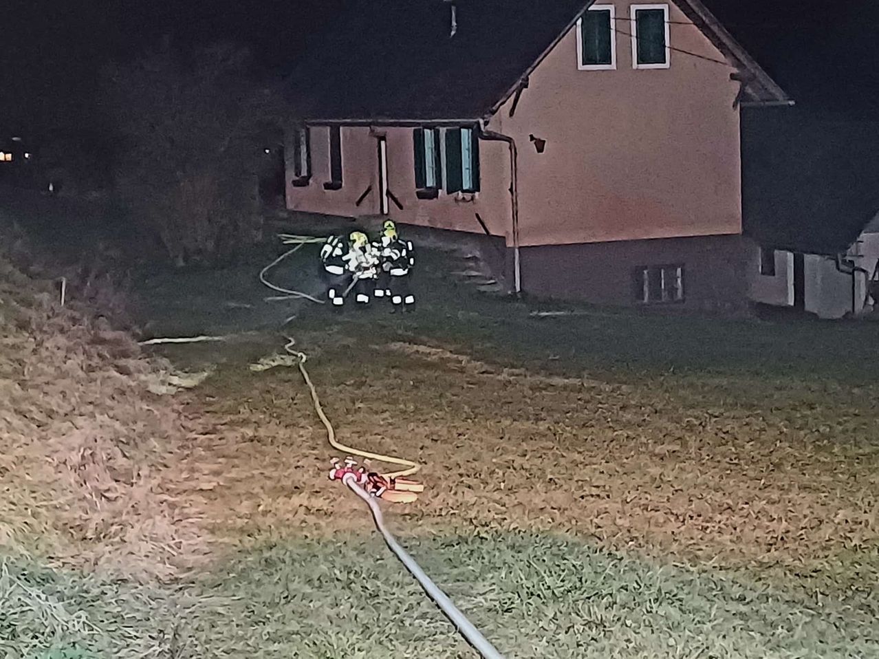 Feuerwehrleute in gelben Uniformen arbeiten bei Nacht vor einem Haus. Ein Schlauch ist auf dem Gras sichtbar. Das Haus hat braune Wände und weiße Fenster.