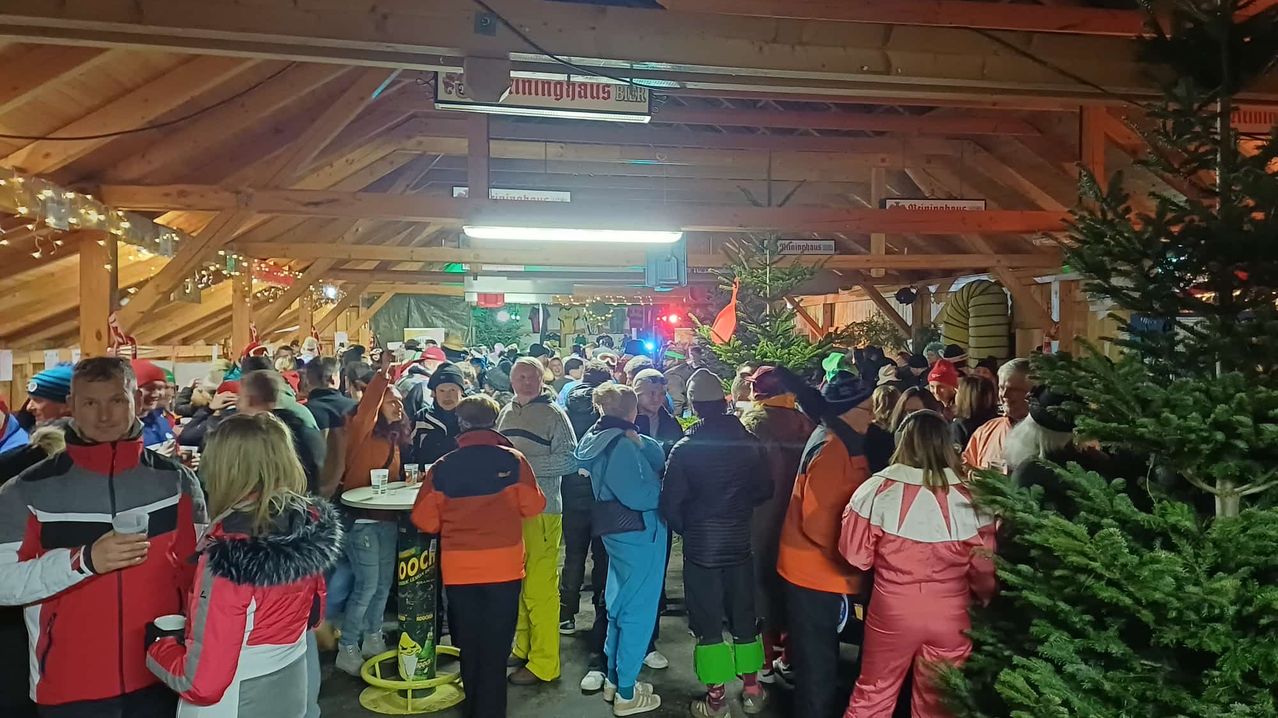 Ein überfüllter Raum mit Holzbalken, vielen Menschen in Winterkleidung, einem Weihnachtsbaum und einem Tisch mit Getränken. Einige Personen tragen Mützen und farbenfrohe Kostüme.