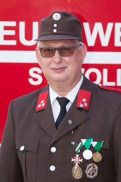 Ein Mann in Uniform mit Medaillen steht vor einem roten Schild mit weißem Text.