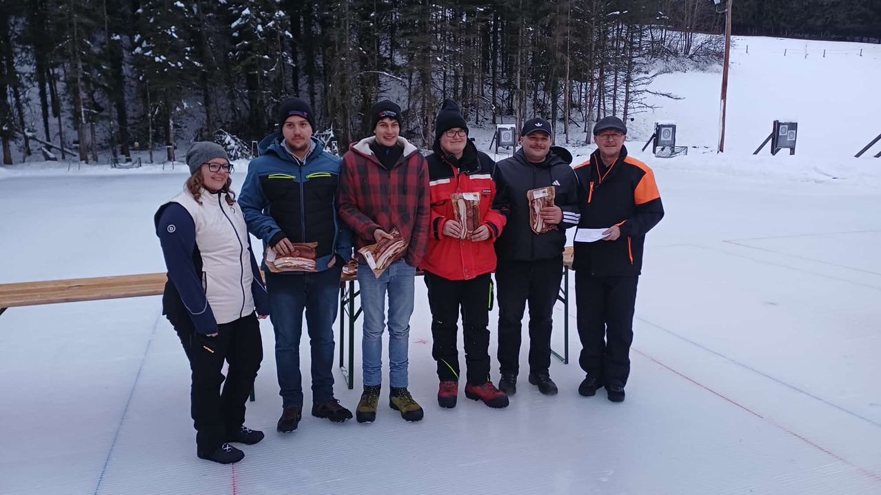 Eine Gruppe von Menschen steht im Schnee, warm gekleidet, hält Stücke Fleisch in den Händen und lächelt für ein Foto. Hinter ihnen sind schneebedeckte Bäume und ein klarer Himmel.