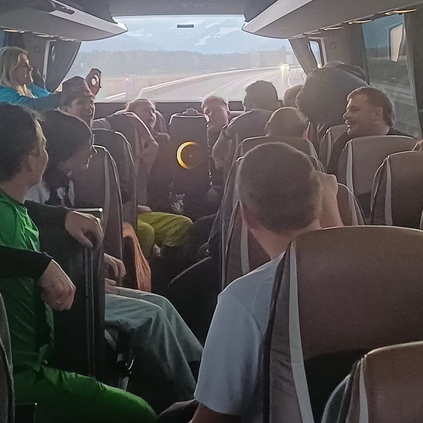 Die Passagiere sitzen in einem Bus; einige lächeln und machen Fotos. Das vordere Fenster zeigt eine Straße und Berge. Die Sitze sind beige, und einer hat einen gelben Kreis darauf.