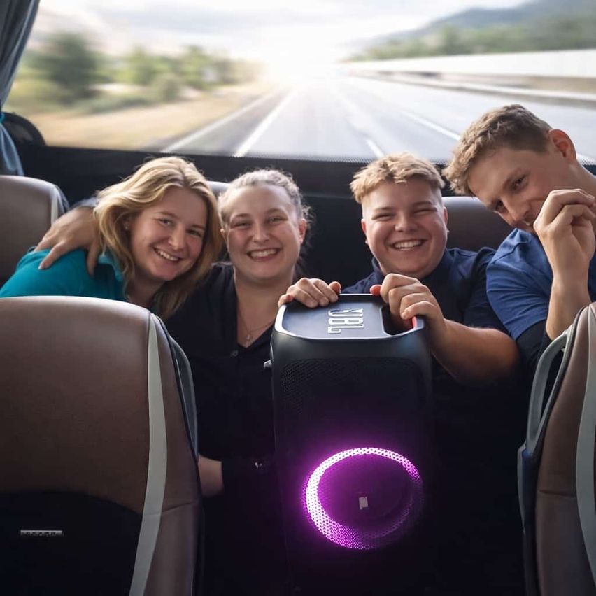 Drei Personen in einem Auto mit einem JBL-Lautsprecher in der Mitte, lächelnd mit der Straße dahinter.