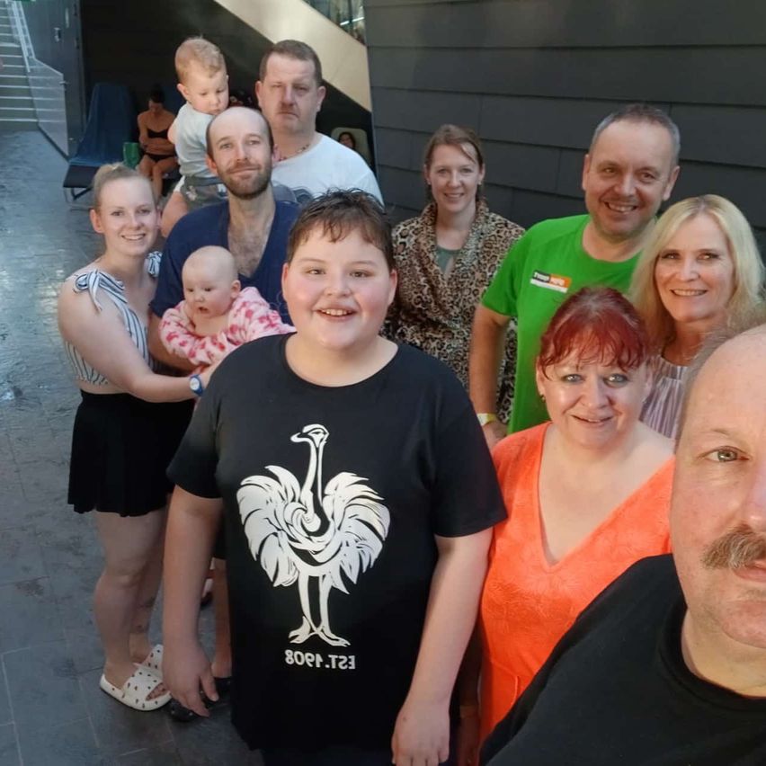 Eine Gruppe von Erwachsenen und Kindern posiert für ein Foto. Die Erwachsenen lächeln und einige von ihnen halten Babys. Der Mann vorne trägt ein schwarzes T-Shirt mit einem weißen Hahn-Design.