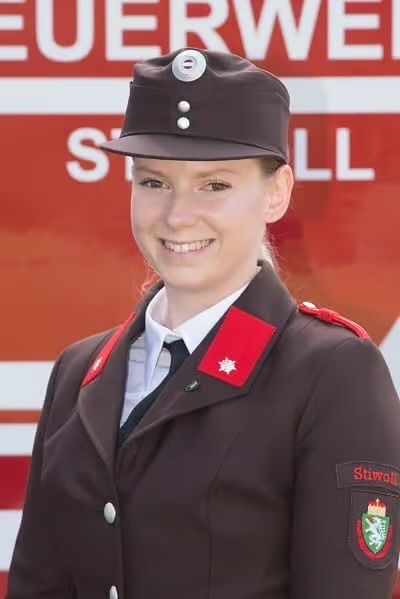 Eine Frau in einer braunen Uniform mit roten Abzeichen und einem Hut steht vor einem roten Schild. Sie lächelt für ein Foto.