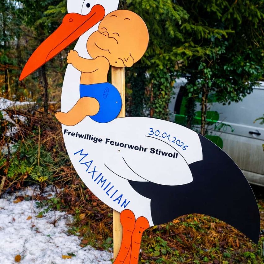 Ein Storch-Schild mit einem Baby darauf steht 'Freiwillige Feuerwehr Stiwol 30.01.2026'. Der Storch ist weiß und schwarz, mit einem orangefarbenen Schnabel und Beinen. Der Hintergrund ist ein verschneiter Bereich mit Bäumen.