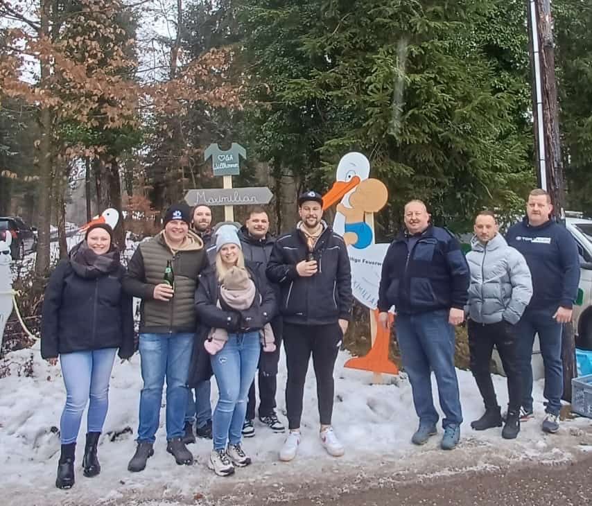 Eine Gruppe von Menschen steht im Schnee, lächelt und hält Getränke in der Hand. Hinter ihnen stehen Bäume und ein großer orangefarbener Storch. Sie sind warm in Winterkleidung gekleidet.