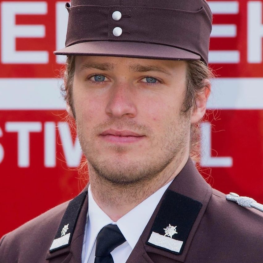 Ein Feuerwehrmann in einer braunen Uniform mit weißen Knöpfen auf seinem Hut steht vor einem roten Schild.