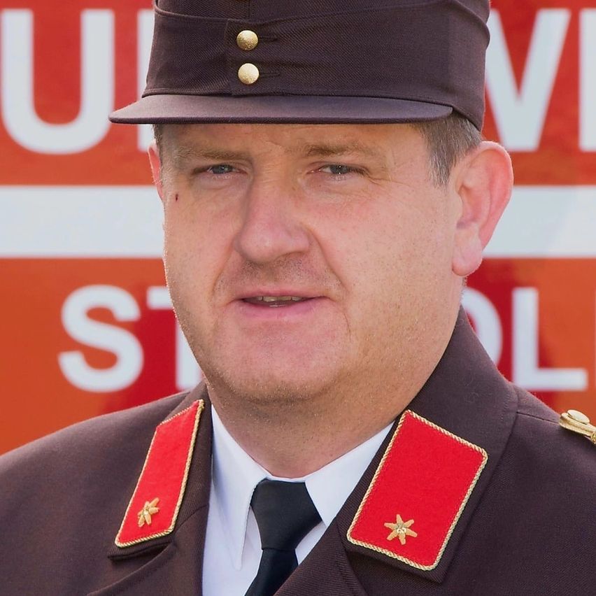 Ein Mann in Uniform steht vor einem roten Schild mit weißen Buchstaben. Er trägt einen braunen Hut mit goldenen Knöpfen und roten Schulterklappen. Sein Gesichtsausdruck ist neutral.