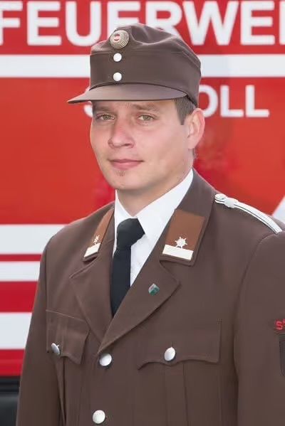 Ein Mann in einer braunen Uniform, Hut und Krawatte steht vor einem Feuerwehrauto. Die Uniform hat einen Abzeichen und Knöpfe. Das Auto hat einen roten und weißen Körper mit dem Wort 'Feuerwehr' darauf geschrieben.