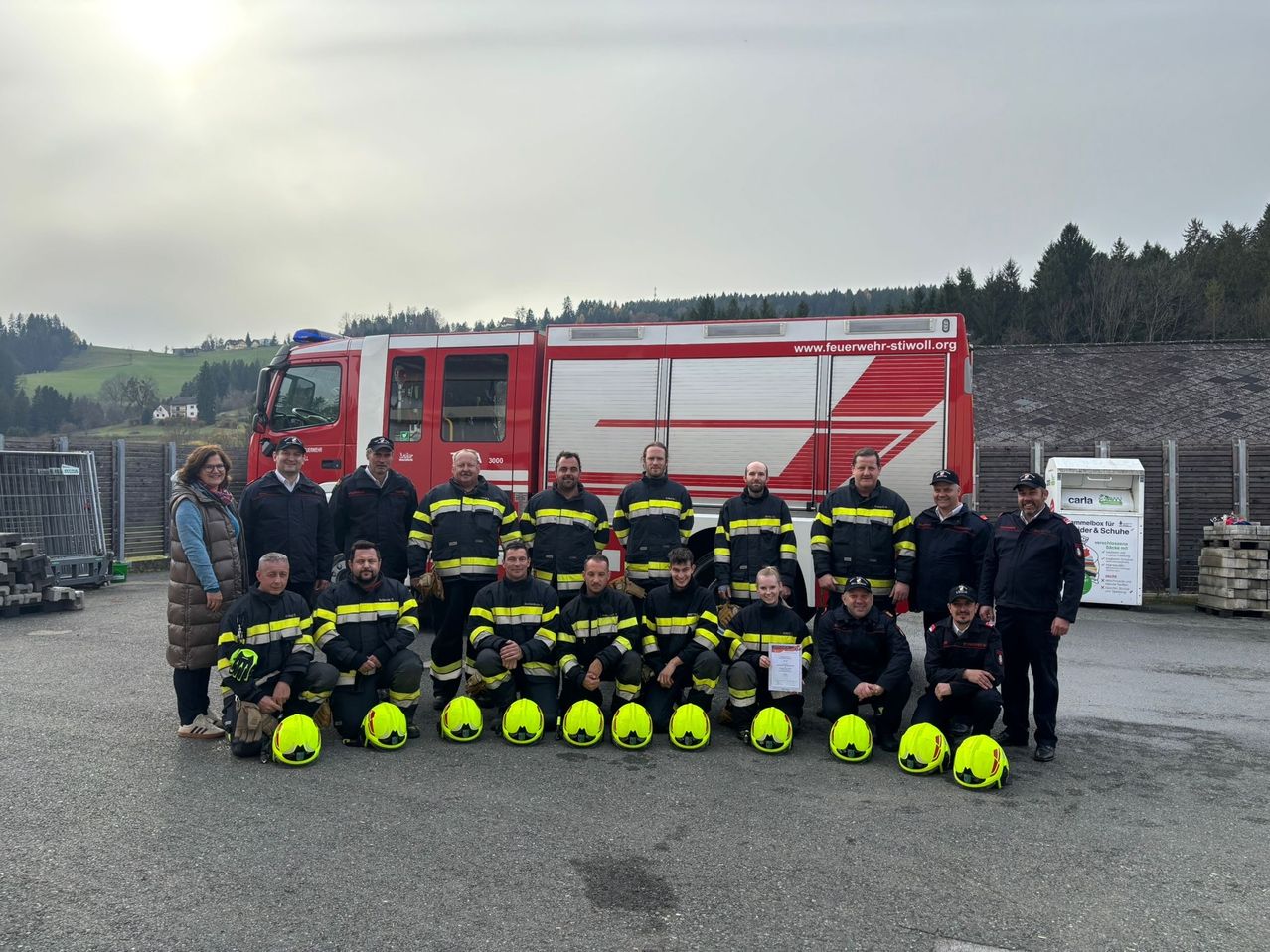 A group of firefighters stands in front of a fire truck, posing for a photo. They wear uniforms with yellow and black stripes. The scene is outdoors, with a building and trees in the background.
