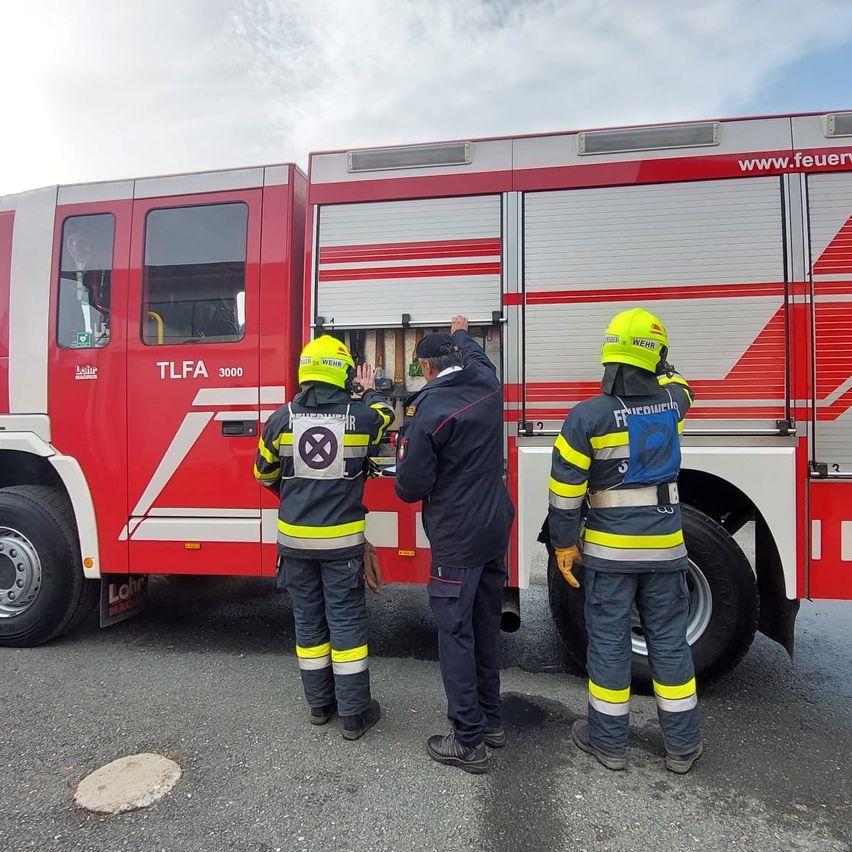 Three firefighters stand in front of a red fire truck. One opens the truck's door while another points inside. They wear yellow helmets and uniforms.