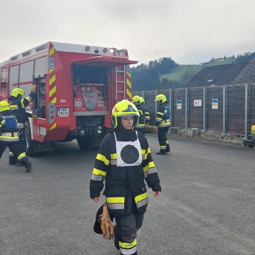 A firefighter walks past a red fire truck. Several firefighters are standing near the truck, preparing for an emergency. Behind them, a wooden fence separates the area from a distant hillside.
