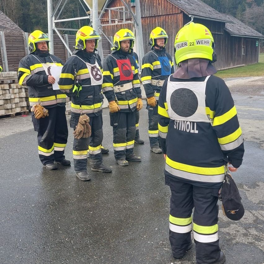 Five firefighters in yellow helmets stand outside, possibly in a training session. One holds a paper. Behind them is a building with a wooden fence.