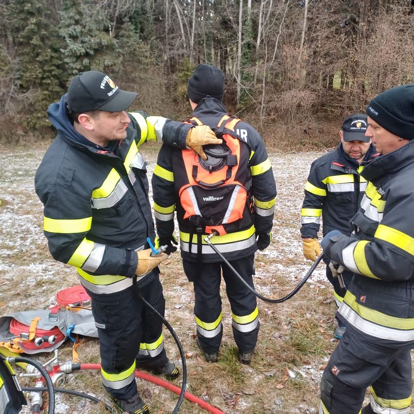 Vier Feuerwehrleute in Schutzkleidung stehen in einem verschneiten Außenbereich. Einer justiert die Ausrüstung eines anderen. Sie sind von Feuerwehrausrüstung umgeben.