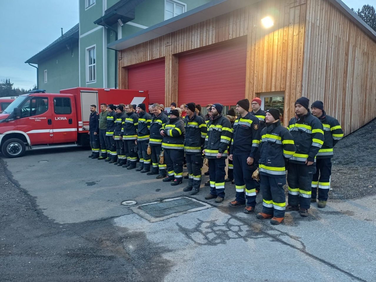 Eine Reihe von Feuerwehrleuten steht vor einer Feuerwache. Sie tragen Uniformen mit reflektierenden Streifen. Ein Feuerwehrwagen ist in der Nähe geparkt.