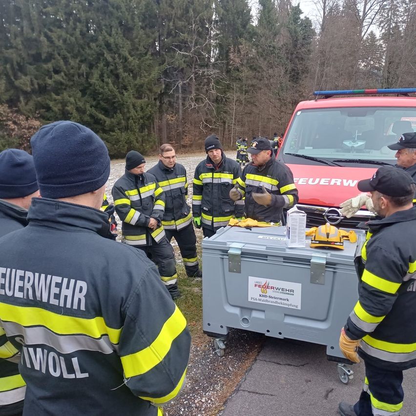 Eine Gruppe von Feuerwehrleuten steht um ein rotes Fahrzeug und einen grauen Container herum, möglicherweise bei einer Übung. Sie sind in Feuerwehruniformen gekleidet.