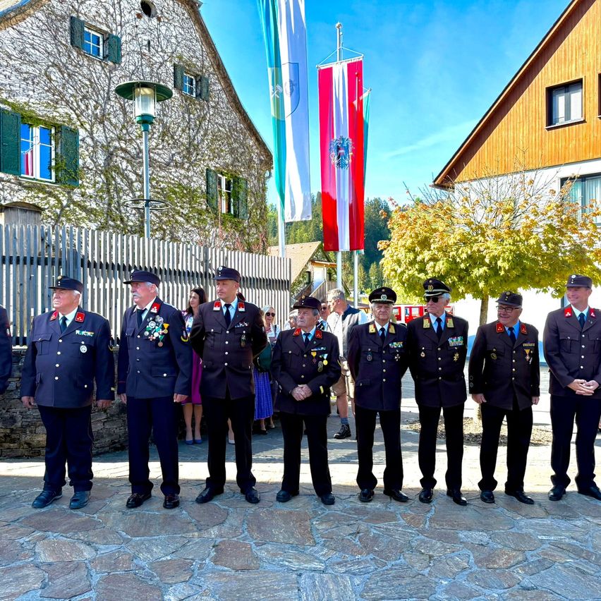 Eine Gruppe uniformierter Männer, möglicherweise Militäroffiziere, steht in einer Reihe vor einem Gebäude, mit Fahnen und Bäumen im Hintergrund.