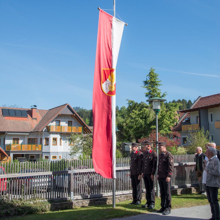 Eine rote Fahne mit Logo ist an einem Mast gehisst. Vier Männer in Uniform stehen davor. Ein Zaun und Bäume sind im Hintergrund. Häuser sind sichtbar, mit Solarpanelen auf den Dächern.