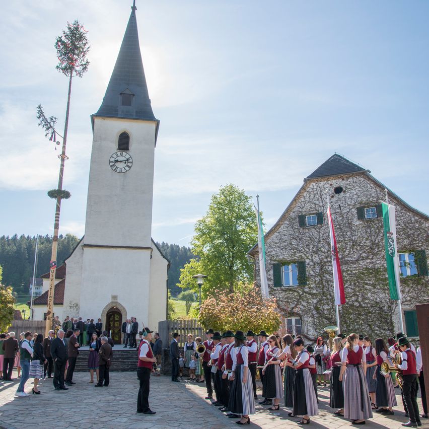 Eine Gruppe von Menschen in traditioneller Kleidung steht vor einer Kirche, mit Flaggen und einem Baum in der Nähe. Eine Uhr ist auf dem Turm sichtbar.