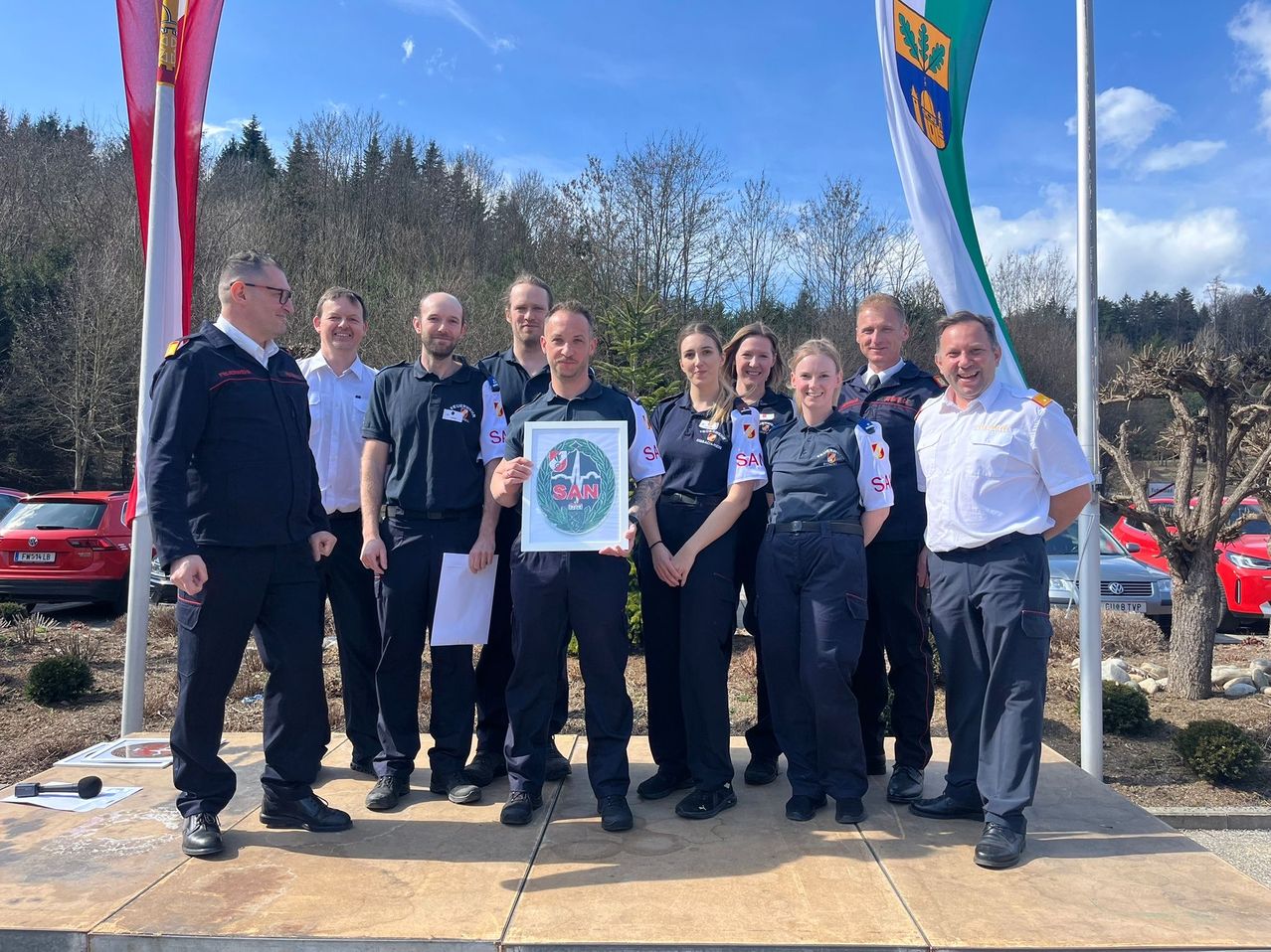 Eine Gruppe uniformierter Personen, wahrscheinlich Feuerwehrleute, steht auf einer Plattform und posiert für ein Foto. Sie halten ein gerahmtes Zertifikat. Hinter ihnen befinden sich Flaggen und Bäume.