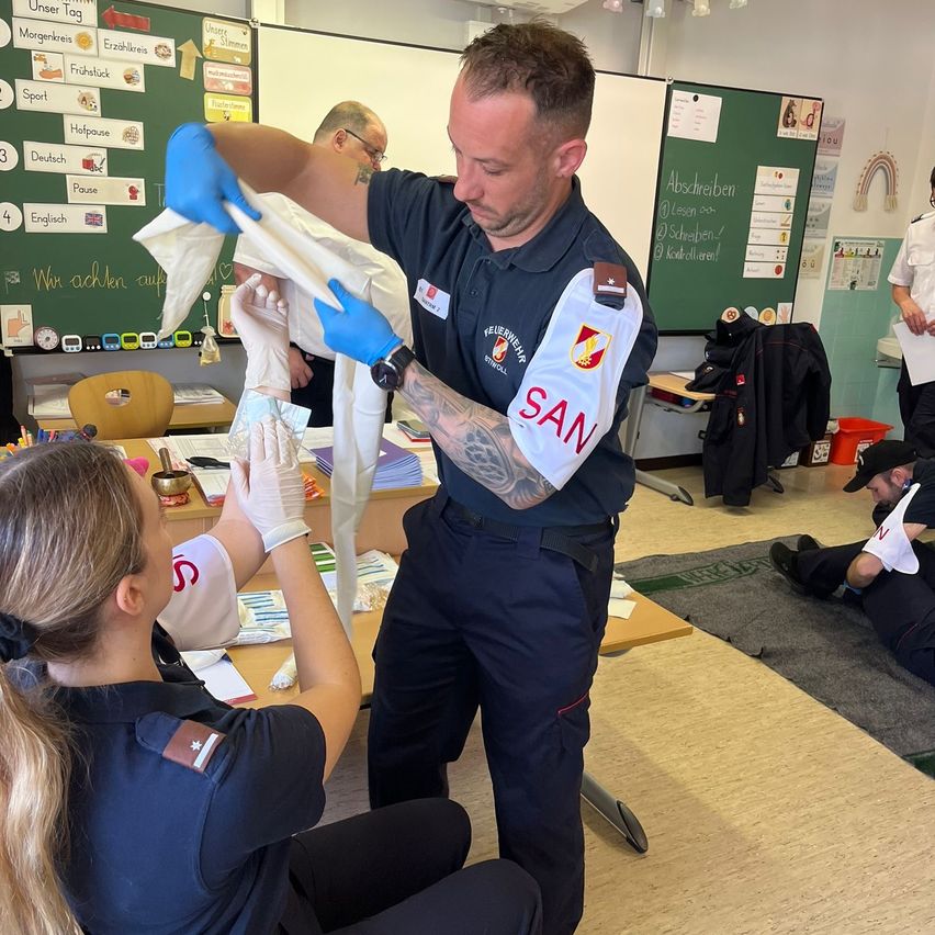 Ein Feuerwehrmann demonstriert einer Frau in einem Klassenzimmer einen Verband. Der Raum hat eine grüne Tafel mit in Deutsch geschriebenen Worten und einen Schreibtisch.
