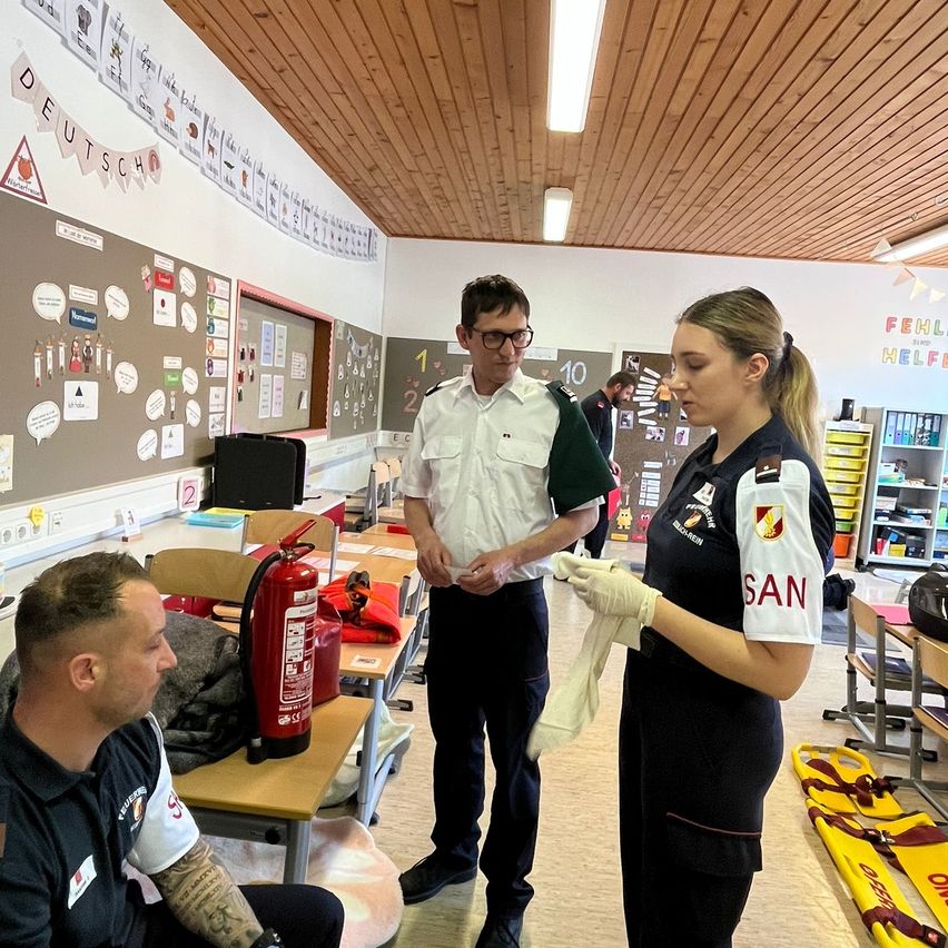 Drei Personen stehen in einem Klassenzimmer mit einem roten Feuerlöscher, einer Bank und einer Wand mit Plakaten. Ein Mann sitzt auf einem Stuhl mit einem bandagierten Arm.