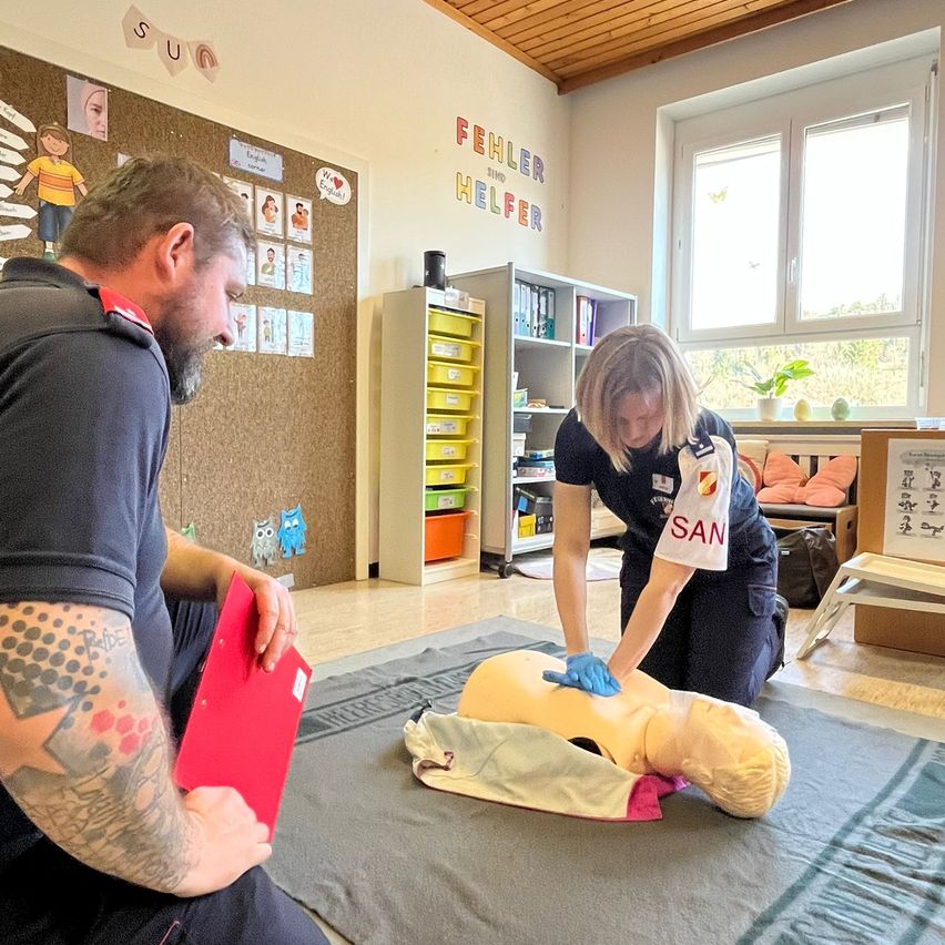 Ein Mann und eine Frau üben CPR an einem Dummies in einem Klassenzimmer. Der Mann hält eine rote Mappe, und die Frau trägt ein SAN-Shirt. Ein Whiteboard befindet sich hinter ihnen mit pädagogischen Postern.