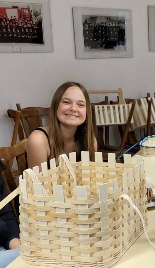 Ein junges Mädchen sitzt an einem Tisch mit einem teilweise fertiggestellten Holzkorb und lächelt in die Kamera. Hinter ihr sind mehrere Holzstühle aufgestellt, und zwei gerahmte Bilder schmücken die Wand.