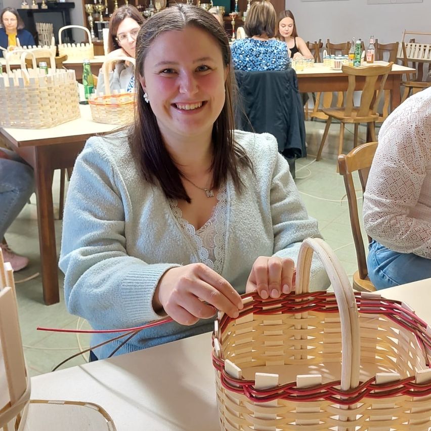 Eine Frau sitzt an einem Tisch und lächelt, während sie einen Korb flicht. Körbe stehen auf dem Tisch und Menschen sitzen im Hintergrund am Tisch.