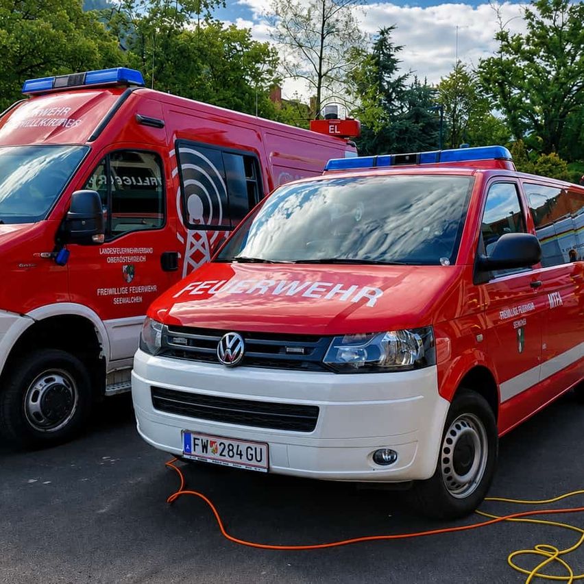 Zwei rote Feuerwehrfahrzeuge sind am Straßenrand geparkt, eines mit dem Wort 'Feuerwehr' auf der Vorderseite und das andere mit dem Wort 'EL' auf der Oberseite.