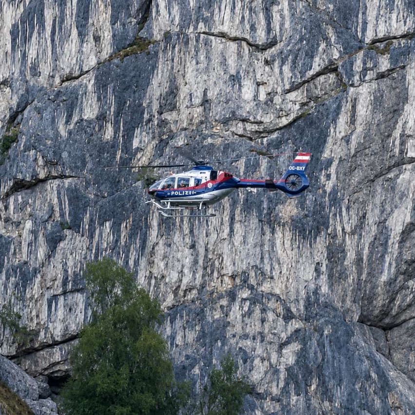 Ein Polizeihubschrauber mit der Registrierung OE-EXY fliegt in der Nähe einer steilen Felswand unter klarem Himmel.