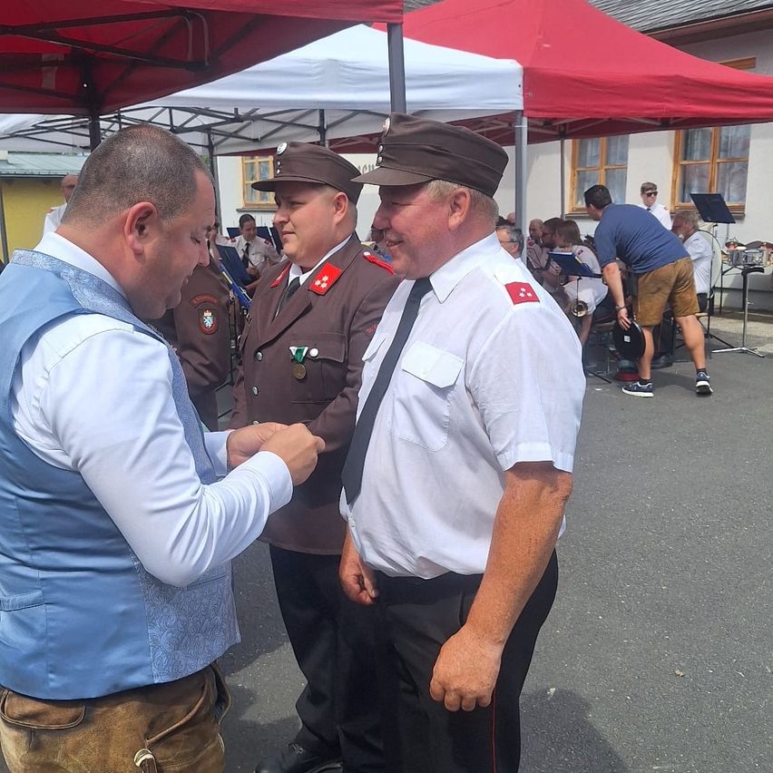 Drei Männer in Uniformen und Hüten stehen vor einem Zelt, einer von ihnen schüttelt einem Mann in einer blauen Weste die Hand. Hinter ihnen spielt eine Band auf Instrumenten.