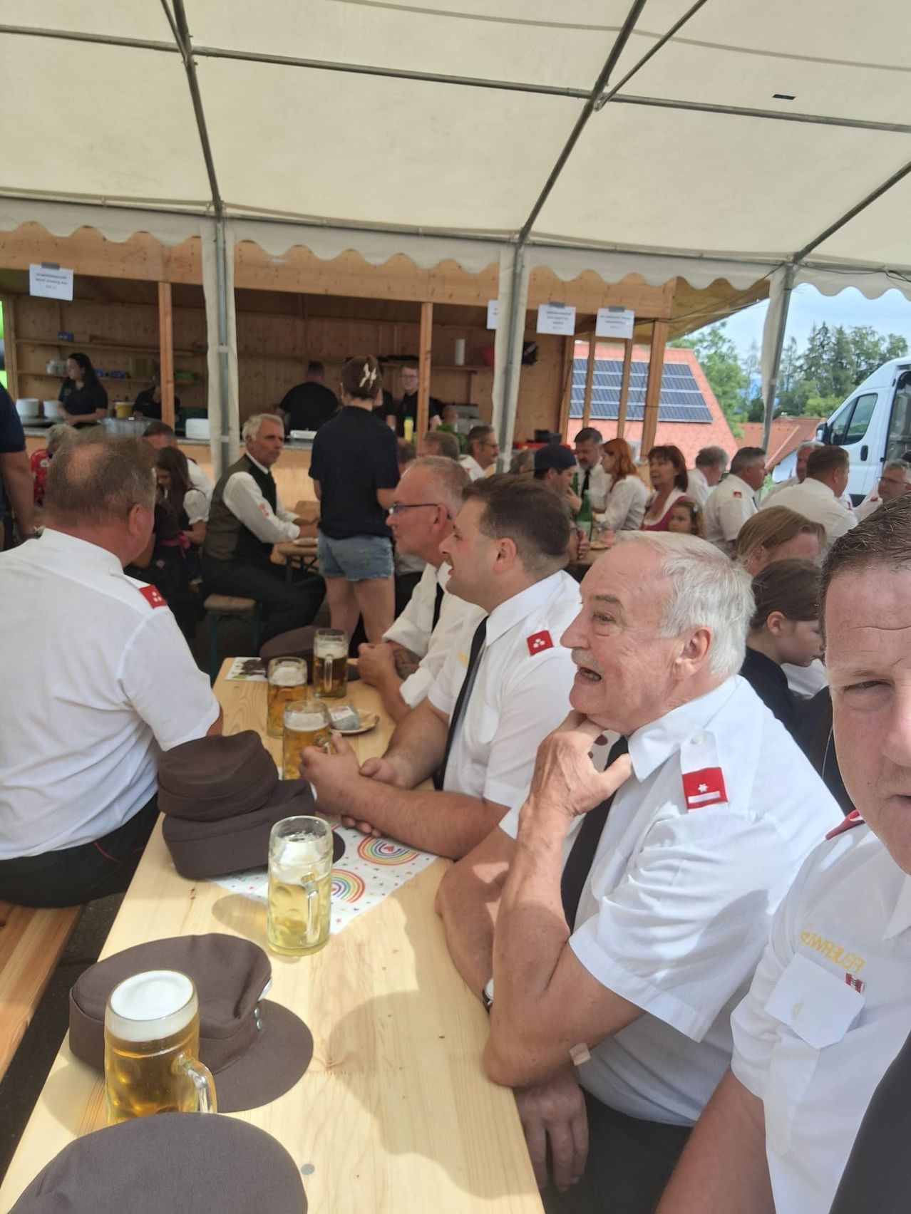 Eine Gruppe von Menschen in weißen Uniformen sitzt an einem Tisch mit Biergläsern und Hüten unter einem Zelt.