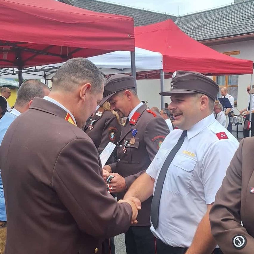 Zwei Männer in Militäruniformen schütteln sich die Hände bei einer Veranstaltung im Freien. Der Mann auf der linken Seite hält einen Stift und Papier. Hinter ihnen befinden sich Zelte, ein Gebäude und andere Menschen.