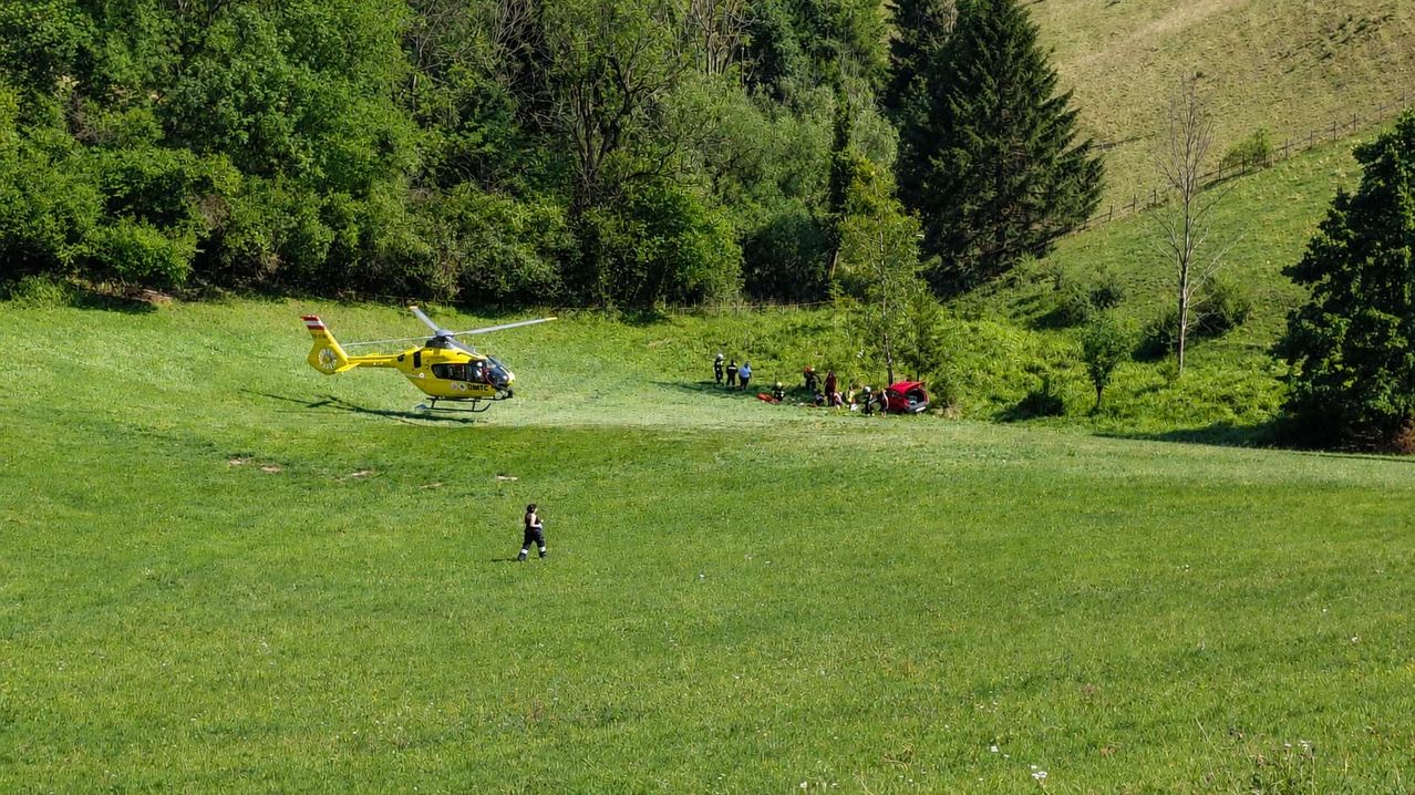 Ein gelber Hubschrauber landet auf einem Grasfeld. Mehrere Menschen sind um ein rotes Auto versammelt. Einige tragen Helme und scheinen medizinisches Personal zu sein.