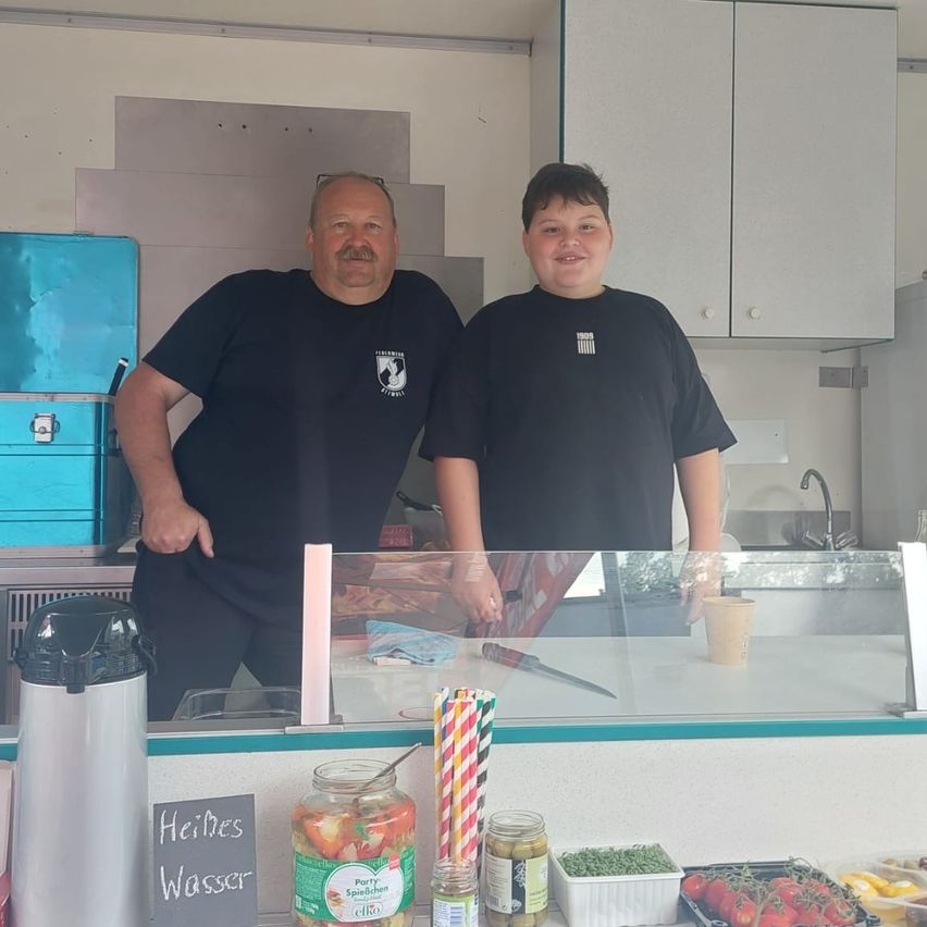 Zwei Männer in schwarzen T-Shirts stehen lächelnd hinter einer Theke in einer Küche. Die Theke hat eine Glasvitrine mit Gläsern, ein Kreidetafelschild mit der Aufschrift 'Heisses Wasser' und einen Kaffee-Thermos.