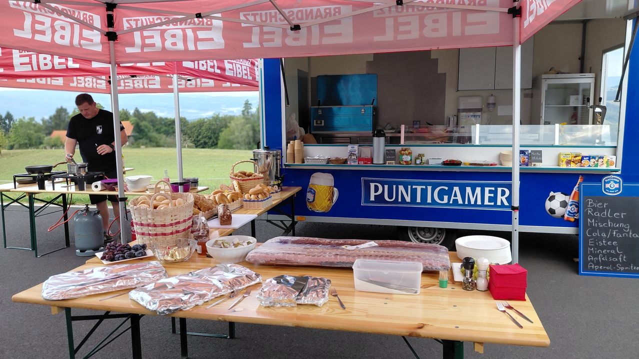 Ein mit Puntigamel beschrifteter Imbisswagen steht auf einer Wiese, mit einem Tisch davor. Der Tisch ist mit verschiedenen Lebensmitteln wie Brot, Fleisch und Gewürzen bestückt. Der Imbisswagen hat ein blau-weißes Farbschema.