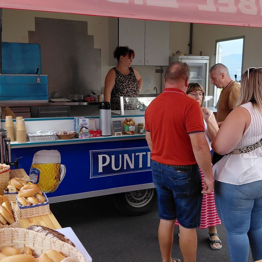 Ein Imbisswagen mit dem Wort Punt an der Seite, eine Frau steht hinter der Theke und mehrere Leute stehen davor, möglicherweise um Essen zu kaufen.