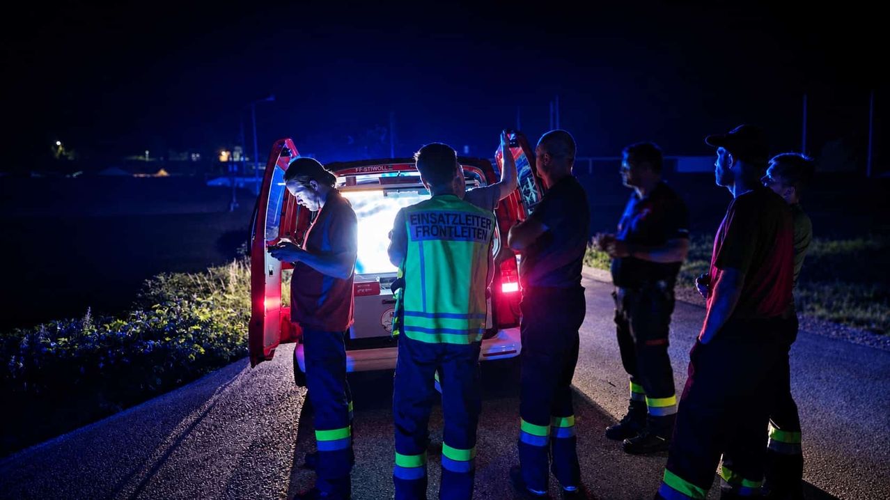 Rettungskräfte stehen um ein Fahrzeug mit offener Heckklappe herum, einer von ihnen hält eine Taschenlampe. Sie sind in reflektierenden Westen und Uniformen gekleidet.