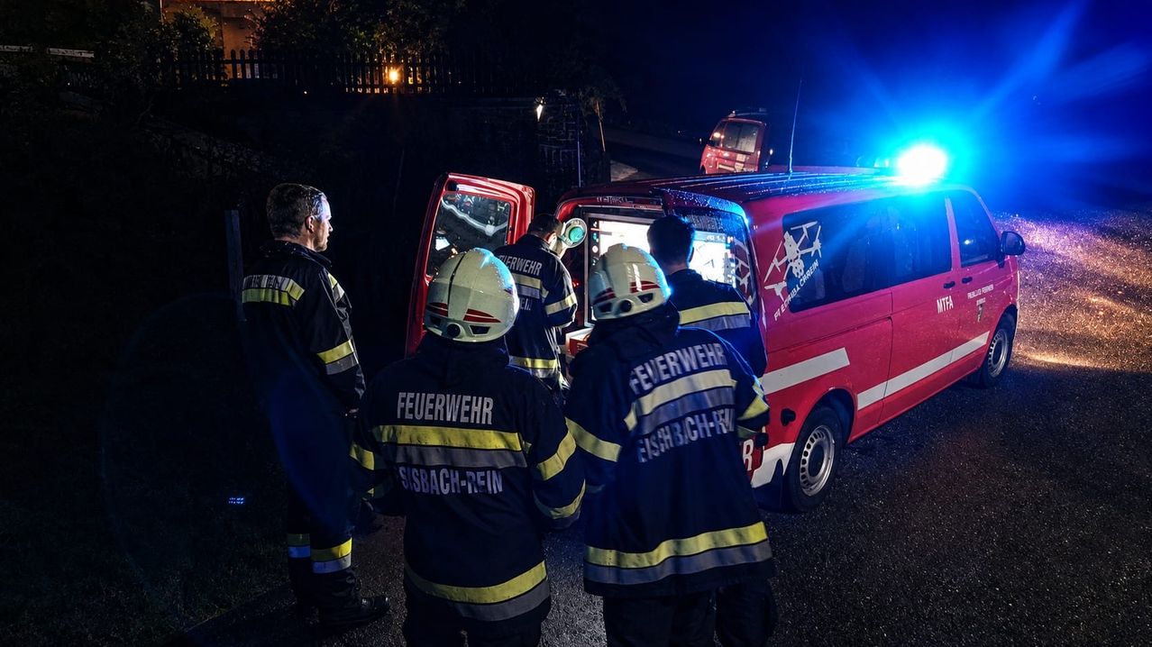 Feuerwehrleute in voller Montur stehen um einen roten Feuerwehrwagen mit offener Tür, bei Nacht mit blinkenden Lichtern.