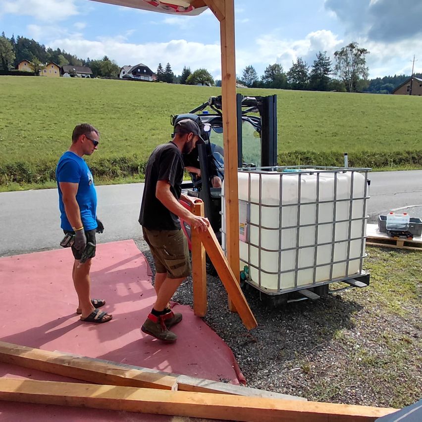 Zwei Männer arbeiten auf einer Baustelle, einer hält eine Holzplanke, während der andere einen Gabelstapler mit einem großen Container bedient. Im Hintergrund sind ein Feld und Häuser zu sehen.