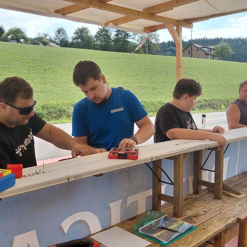 Drei Männer arbeiten unter einem hölzernen Vordach an einem langen weißen Brett. Sie sind von Werkzeugen und Papieren umgeben. Im Hintergrund befindet sich ein grünes Feld und ein Haus.
