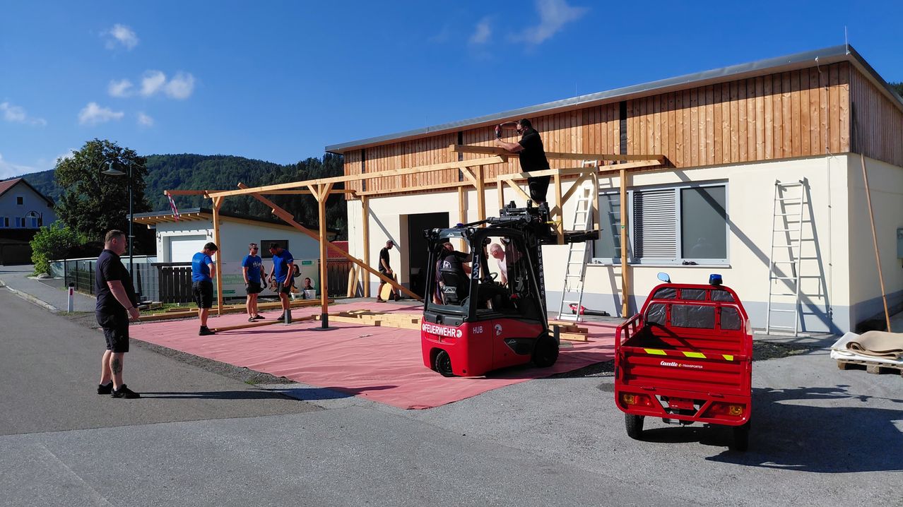 Eine Gruppe von Menschen arbeitet an einer Holzkonstruktion vor einem Gebäude. Ein Gabelstapler steht in der Nähe und wird von einer Person bedient. Eine Leiter ist an das Gebäude gelehnt. Der Himmel ist klar und blau.
