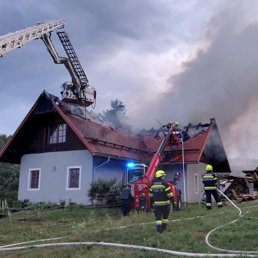 Feuerwehrleute bekämpfen einen Hausbrand mit einem Drehleiterwagen und Schläuchen. Rauch steigt vom Dach auf.