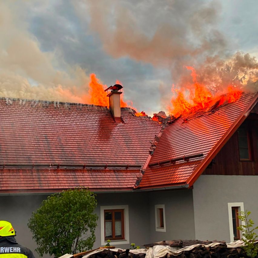 Ein Haus mit einem roten Dach steht in Flammen, wobei die Dächer von den Flammen umhüllt sind und Rauch in den Himmel aufsteigt. Ein Feuerwehrmann steht in der Nähe, möglicherweise zur Beurteilung der Situation.